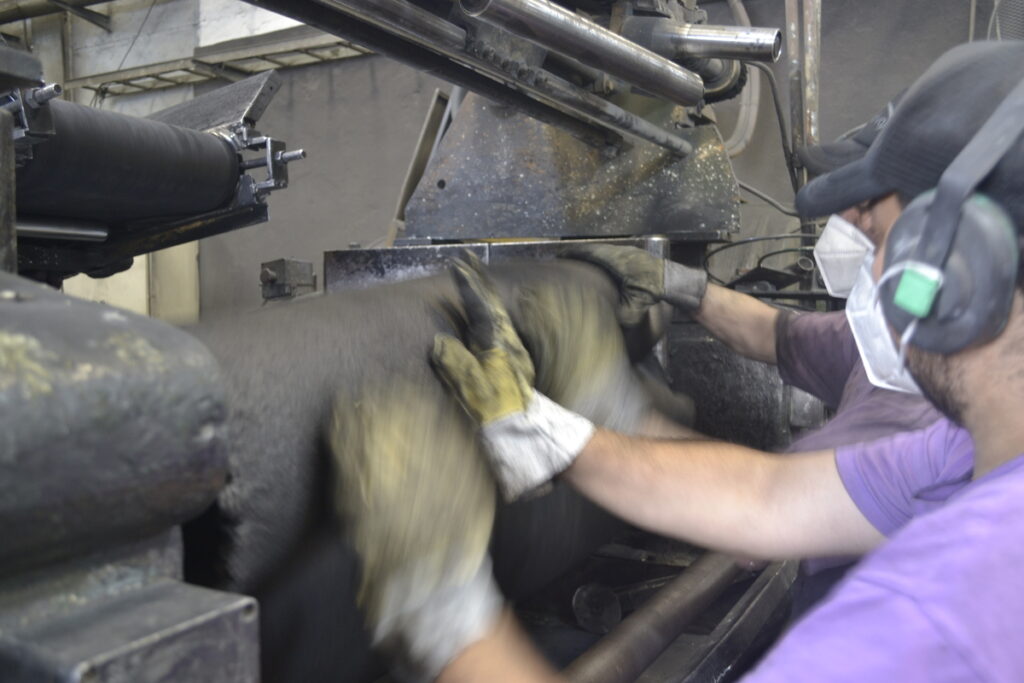 Operarios con guantes, mascarillas y protectores auditivos manipulando caucho en máquina mezcladora industrial